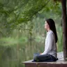 Woman meditating cross-legged on a wooden deck by a pond (illustrative photo)