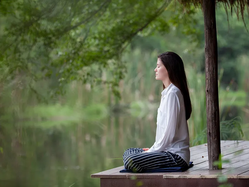 Woman meditating cross-legged on a wooden deck by a pond (illustrative photo)